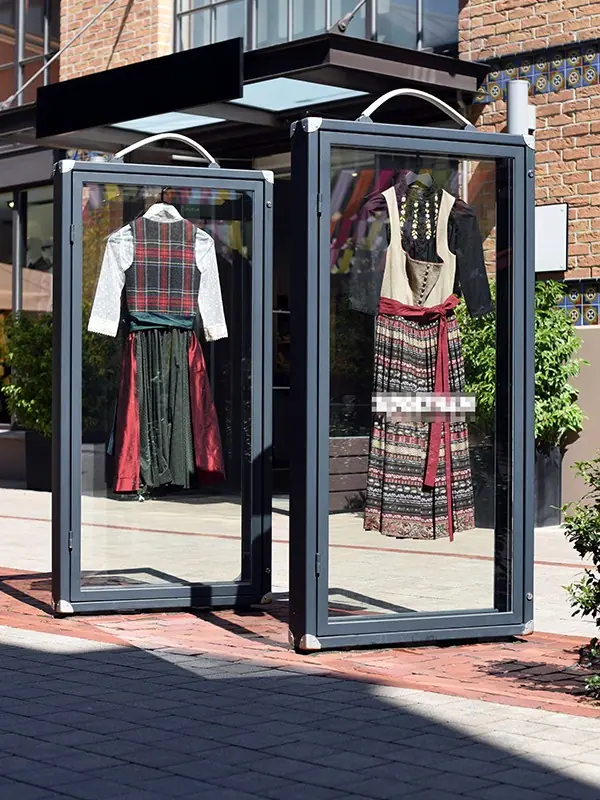 Zwei Displaykabinen mit Rahmen aus Metall und Glas, darin Trachtenkleider, auf einem Platz